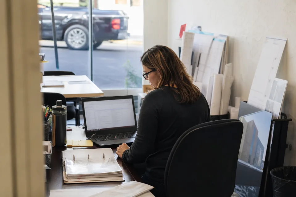 Project manager at a small general contractor office working through a spec binder with a laptop