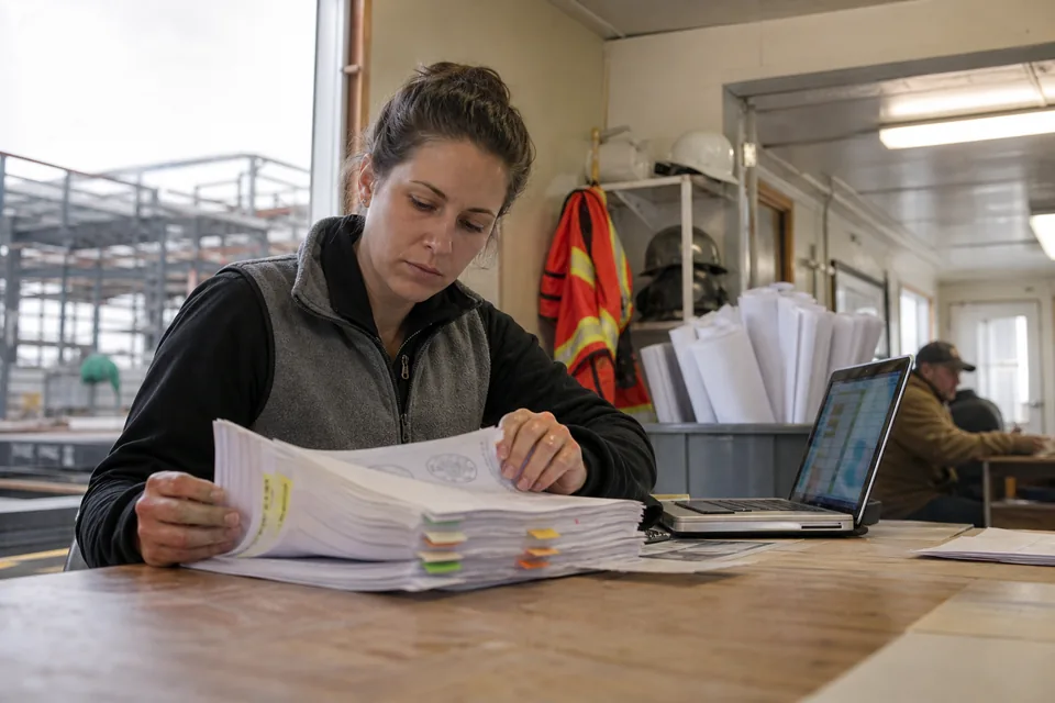 Project engineer in a site trailer sorting tabbed submittal packages with stamp marks next to a laptop showing a register grid