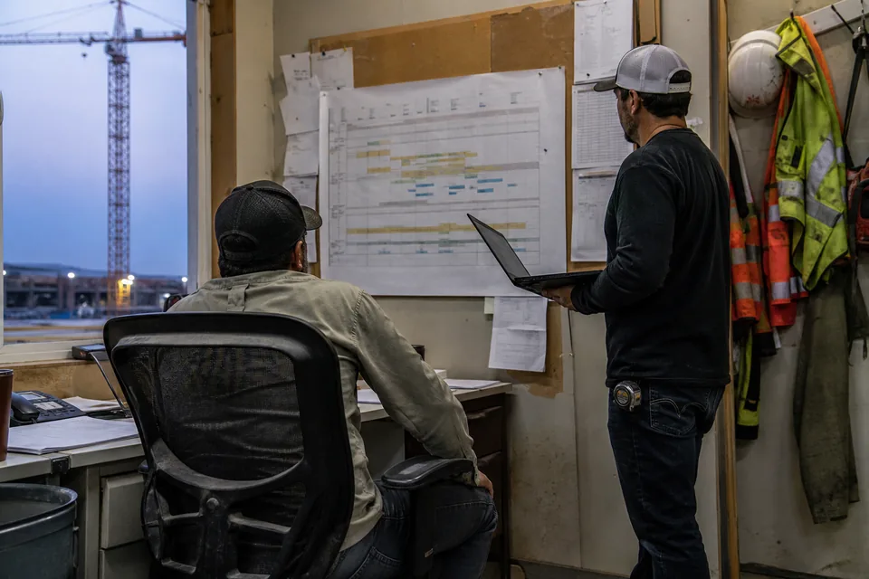 General contractor project executive and project engineer reviewing a lookahead schedule beside a submittal list in a field office