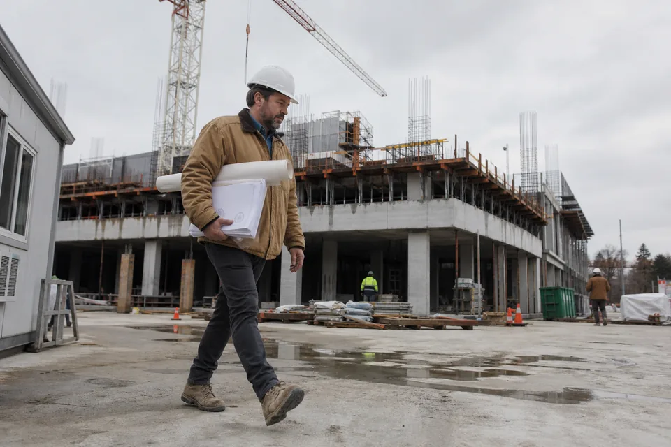Superintendent in a white hard hat walking across a concrete jobsite with a tower crane and a mid-rise structure behind, carrying a rolled drawing set and a stamped submittal package