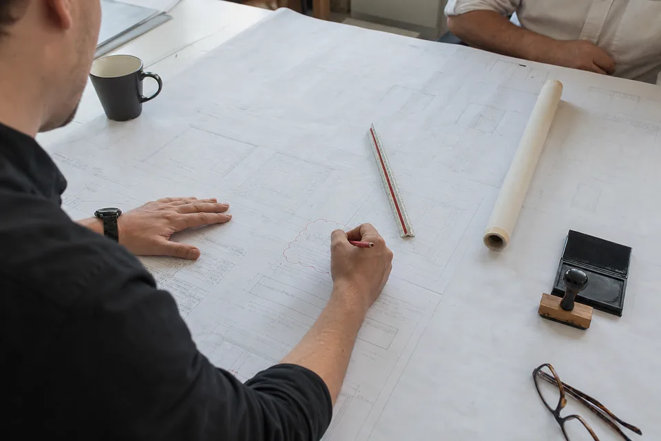 Architect marking up a large shop drawing set with a red pencil on a wide table, scale ruler and stamp beside the sheets