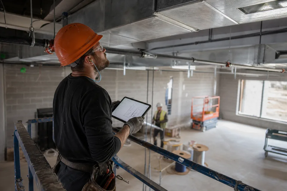 Mechanical subcontractor on a rolling scaffold checking a duct shop drawing on a tablet against installed ductwork at an open ceiling
