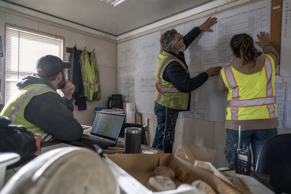 General contractor project team in a site trailer pinning a printed submittal schedule next to drawings on the wall