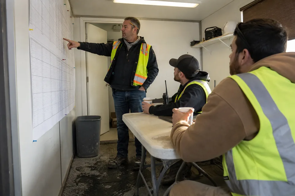 Superintendent pointing at a printed submittal log taped to the trailer wall during a coordination meeting with two foremen