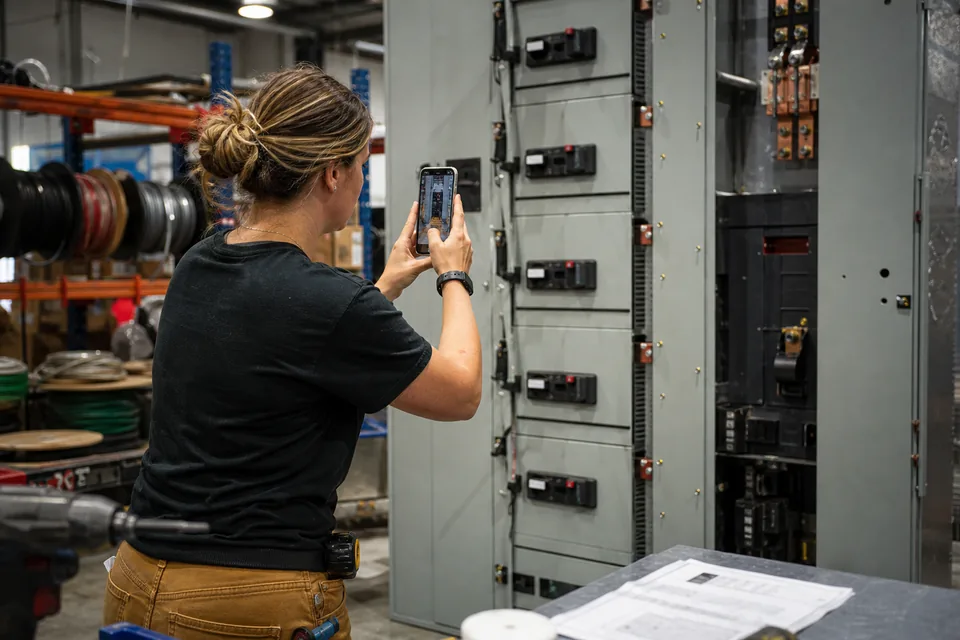 Electrical subcontractor project manager photographing a switchgear panel for a submittal package in a prefab shop